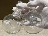 Two round bottom glass lab beakers held by a gloved hand, showing brand markings, ground glass joints, and 250 ml volume indicators.