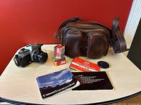 Overview photo showing Canon AT-1 camera, unopened film boxes, manuals, and the vintage leather camera bag on a table against a red background.