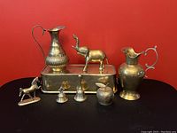 Photo of the entire brass collection arranged against a red background, showing pitchers, figurines, planter, bells, and apple-shaped item.
