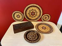Six vintage carved wooden decor items arranged on a light wood table against red wall: five nesting round plates with floral and geometric patterns and one rectangular bird-carved jewelry box