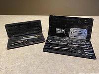 Both vintage Wild Heerbrugg drafting sets displayed side by side on surface, showing all included drafting tools in black cases.