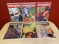 Six Snotgirl comic books displayed in two rows on a table against a red wall, showing front covers of issues 3 through 8 with colorful artwork and characters.
