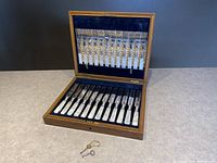 Open wooden box lined with dark blue velvet showing 12 knives in lower compartment and 12 forks in upper lid, two winding keys resting in front