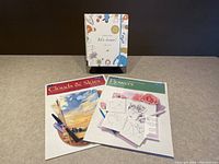 Front view of three art instruction items on a table: two instructional art books and a hardcover sketch pad kit.