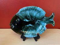 Blue Mountain Pottery ceramic fish shaped dish displayed upright on stand against red background showing glaze and colors.