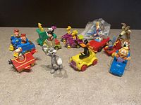 All toy figures of Looney Tunes and Peanuts characters arranged on a surface, showing 12 different toys in their various cars and forms.