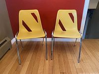 Front view of two yellow Mid Century Modern stacking chairs with plastic seats and metal legs against a red wall.