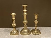 Three brass vintage candlesticks with different shaped bases and heights on plain gray surface with brown background.