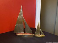 Two vintage brass sailboats displayed on a black surface against a red and gray background. Larger boat features a wide, flat base and two sails; smaller boat has a solid base with two sails, one taller and one smaller triangular.