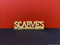 Front view of brass wall-mounted hanger rack spelling 'SCARVES' in uppercase letters against a red background.