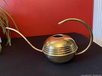 Front and side view of the spherical mid century modern brass watering can with curved spout and handle on a black surface against a red wall, showing its polished finish and ridged body.