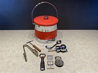 Full lot overview displaying the vintage red and chrome ice bucket with lid, metal ice tongs, wooden muddler, two Labatts branded bottle openers, a silver cocktail jigger, and two decorative metal wall hangings.