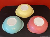 Three nested Pyrex mixing bowls placed to show the outside bottoms, revealing red (faded), yellow (shiny), and turquoise (shiny) colors and handles on each.