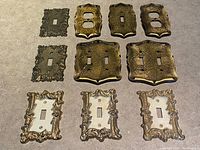 Top-down view of 10 vintage brass and metal switch and outlet plate covers with ornate Victorian designs and patina on a stone surface.