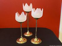 Set of three brass and frosted glass tulip candle holders on a black surface against a red background, showing tiered heights and frosted petal-shaped shades.