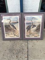 Both framed egret bird pictures shown upright outdoors on pavement, side by side.