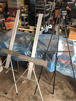 Three art easels shown side by side in a garage setting, two aluminum with white frames and one black wooden easel.