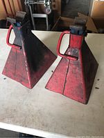 Photo showing two red metal car jack stands from an angled side view on a white table, exhibiting wear and use marks.
