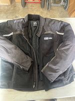 Photo of the front of the black Can-Am Spyder motorcycle jacket with mesh and fabric sections, showing overall condition and embroidered logo on chest.