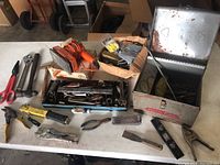 Lot of vintage tools spread on table including toolbox, wrenches, pliers, scissors, hand drill and others.
