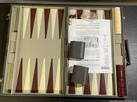 Open backgammon board showing playing surface with maroon and cream triangles, game pieces, dice, dice cups and instructions inside the case.
