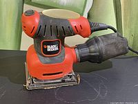 Side view of Black & Decker palm sander showing orange and black body, rubber grip and dust collection bag.