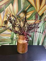 Full view of hammered copper vase with faux plant arrangement inside on black surface against floral green-yellow background.