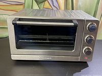 Front view of the stainless steel Cuisinart toaster oven placed on a black surface. The glass door and control knobs are visible.