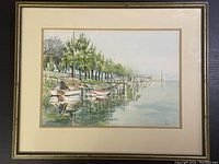 Framed watercolour artwork showing a serene scene of boats moored along a tree-lined dock extending into calm water. The painting features green trees, reflections in water, and softly faded colors.