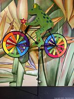 Full view of the metal frog on bike wind spinner showing the colorful pinwheel wheels and the flower basket detail.