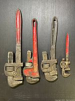 Photo showing all four pipe wrenches of varying sizes with red and black handles, laid flat on a black surface.