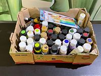 Box containing many small paint bottles and a package of paintbrushes on top