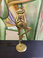 Full view of gold-tone metal frog figurine standing on one leg with round base.