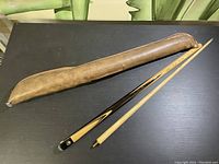 Entire pool cue disassembled in two pieces and brown leather case laid out on a black surface