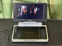 Front view of Panasonic portable DVD player opened, screen showing a movie scene, base with control buttons and AC power cord attached