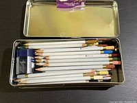 Opened metal tin showing 20+ white-bodied colored art pencils and a metal pencil sharpener inside.