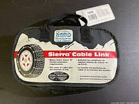 Photo of the front of the black carrying case for Sierra Cable Link tire chains. The label features a tire illustration, the brand name "Sierra Cable Link Chain Company," and indicates compliance with S.A.E. Class "S" clearance requirements.