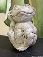 Front view of a small concrete frog garden ornament showing its relaxed pose with hand on cheek.