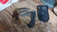 Three locking folding knives on a wooden surface, two with wooden and brass handles, one with black handle, plus black fabric sheath.