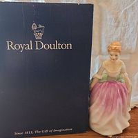 Photo of the Alison Royal Doulton porcelain figurine standing next to the original dark blue box with gold Royal Doulton branding.