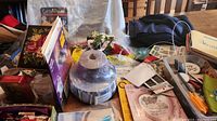 Photo showing a table densely packed with multiple craft items including a purple spool of ribbon, artificial flowers, scissors, booklets, and packaged craft supplies.