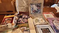 Full view of crafting supplies on table including sealed and boxed needlepoint kits, pattern booklets, skeins of yarn, needles, and hooking materials.