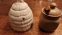 Two ceramic honey pots, one beehive-shaped with bee decoration and one plain round pot with lid and honey dipper.