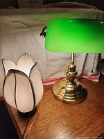 Two lamps on wooden surface against beige fabric. One is a brass banker lamp with green glass shade and pull chain. Other is white flower-shaped glass lamp with black petal outlines.