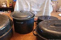 Multiple black speckled roasting pans with lids placed on a table surface under natural indoor lighting.