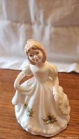 Front view of Royal Doulton porcelain figurine of a young woman in white dress with green floral accents, holding bonnet.