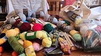 Photo showing a large variety of yarn skeins in multiple colors alongside packaged knitting needles and crochet hooks arranged on a surface