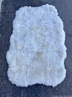 Full view of a white faux fur rug displaying its soft, dense fibers and irregular shape on a dark floor surface.