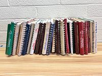 Photo showing multiple coil-bound and hardcover cookbooks arranged vertically on a table showcasing the variety of colors and titles on the spines.