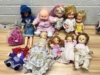 Front view of 10 assorted vintage dolls arranged on a light wood surface against a white brick wall background. Dolls include vinyl and fabric types with varied clothing and hairstyles.